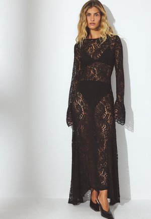 Abito maxi in pizzo nero con maniche lunghe, caratterizzato da intricate fantasie floreali e polsini a campana, sovrapposto a un indumento intimo nero solido.