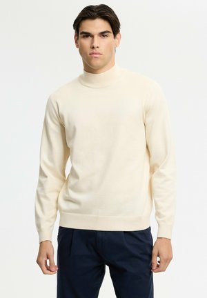 Joven con cabello oscuro que lleva un jersey de cuello alto color crema y pantalones oscuros, de pie frente a un fondo claro y liso.