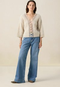 Pull en laine beige avec un décolleté en V profond et des détails en dentelle au centre, associé à un jean bleu clair à jambes larges et des chaussures à motifs.