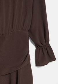 Braune Bluse mit langem Arm, strukturierter Oberfläche, gerafften elastischen Bündchen und gerafftem Nahtdetail an Taille und Ärmelsaum.