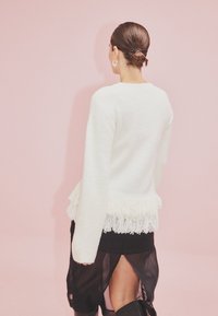 Bela pulover s frufru robom, teksturirano blago in dolgimi rokavi, v kombinaciji z črno krilom z bočnim razporkom, postavljenim na rožnati ozadju.