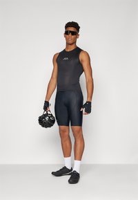 Schwarzes ärmelloses Radtrikot und enganliegende Shorts aus strukturiertem Stoff, Model hält einen schwarzen Helm, kombiniert mit schwarzen Radschuhen und weißen Socken.