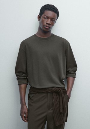 Dunkelgrünes Langarmshirt mit Rundhalsausschnitt, kombiniert mit passender hoch taillierter Hose. Das Shirt hat umgeschlagene Ärmel und ein gebundenes Taillendetail.