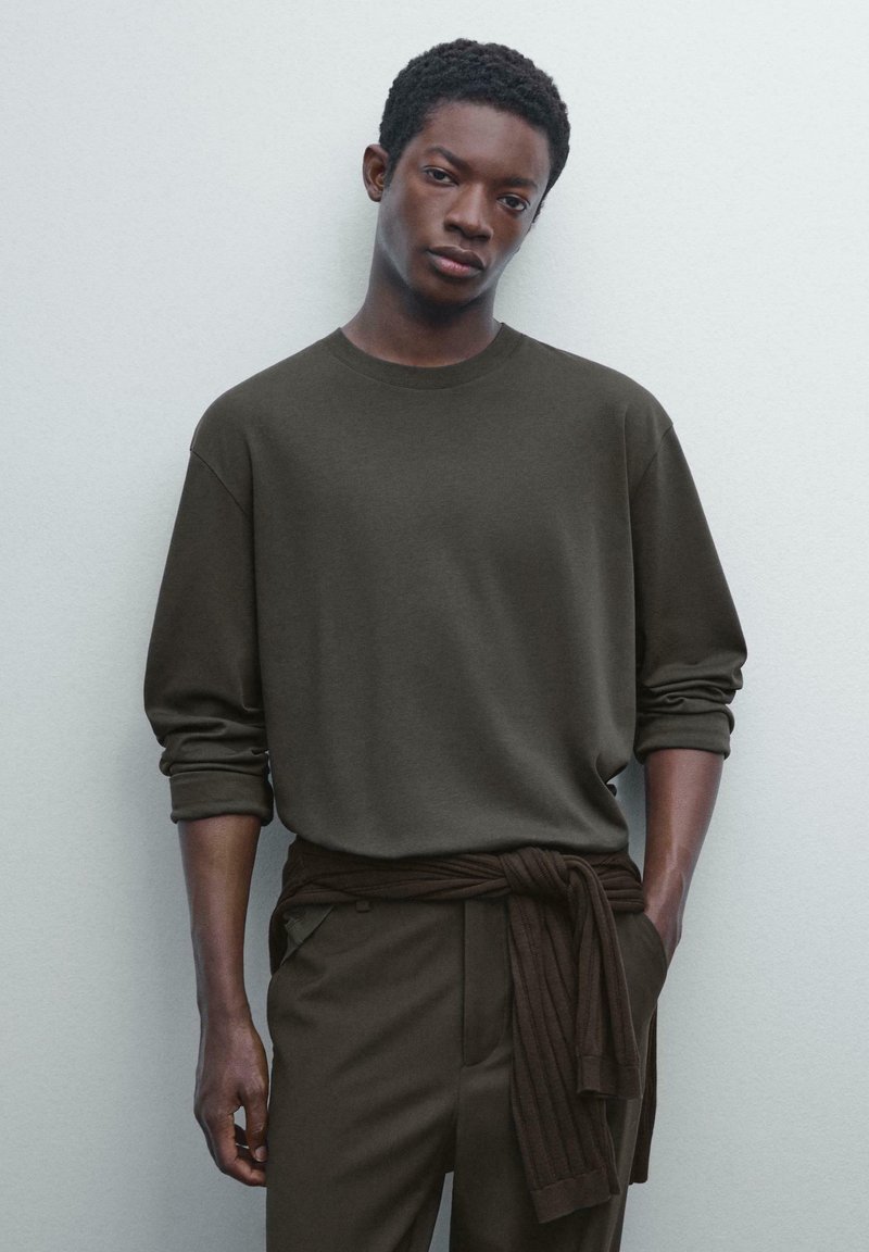 Dunkelgrünes Langarmshirt mit Rundhalsausschnitt, kombiniert mit passender hoch taillierter Hose. Das Shirt hat umgeschlagene Ärmel und ein gebundenes Taillendetail.