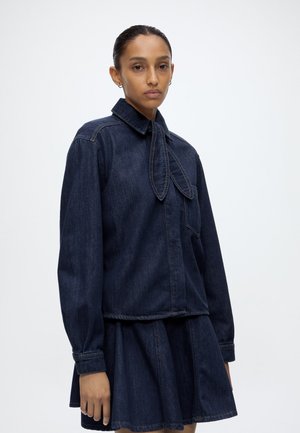 Vrouw met naar achteren getrokken haar, die een donker denimjack draagt met een gestrikte kraag en een bijpassende denimrok tegen een effen achtergrond.