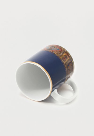 Tasse en céramique blanche avec des motifs cachemire bleus et rouges-dorés complexes, posée sur le côté avec la anse vers la droite sur une surface blanche.
