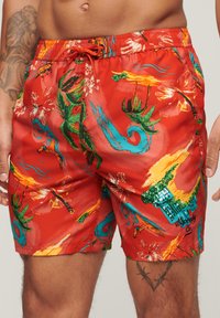 Pantaloni da bagno rossi con stampa tropicale che presenta foglie verdi, onde blu e disegni floreali. Include una vita con cordoncino e una texture liscia.
