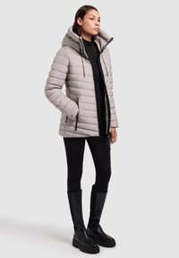 Quilted lys grå pufferjakke med høj krave og justerbar hætte, lynlås foran og to sidelommer. Båret med sorte leggings og støvler.