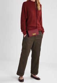 Rode geribbelde gebreide trui, olijfgroene cargobroek met zakken, bordeauxrode crossbodytas met goudkleurige details en donkere platte schoenen.