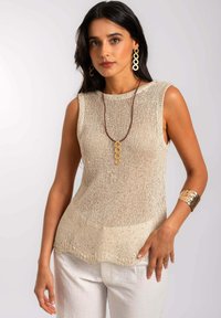 Top sans manches en tricot crème avec une finition texturée, un ourlet arrondi et un léger brillant. Associé à un pantalon en lin blanc et des accessoires dorés.