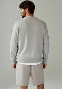 Lysegrå sweatshirt med ribbet krave og manchetter. Har et rektangulært patch på bagsiden, parret med lysegrå shorts.
