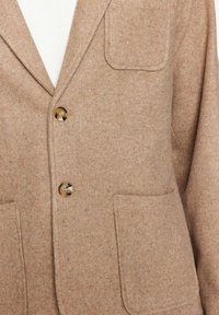 Blazer in lana marrone con due tasche frontali, revers a punta e due bottoni. La texture del tessuto appare morbida e liscia.