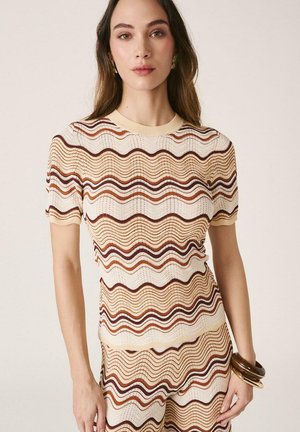 Mujer con conjunto de top y pantalones de manga corta a juego, con un patrón de rayas onduladas en beige, marrón y óxido, adornada con pulseras en la muñeca derecha.