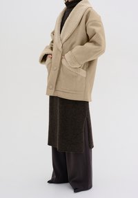 Beige kabát z vlněné směsi s umělou kožešinou, velkým límcem, dvěma předními kapsami a knoflíkovým zapínáním, doplněný tmavohnědými sukněmi a širokými kalhotami.