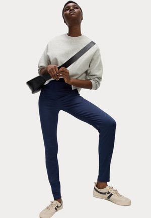 Osoba ubrana w jasnoszary sweter, granatowe legginsy, beżowe sneakersy i czarną torbę na ramię, stojąca z podniesioną jedną nogą.