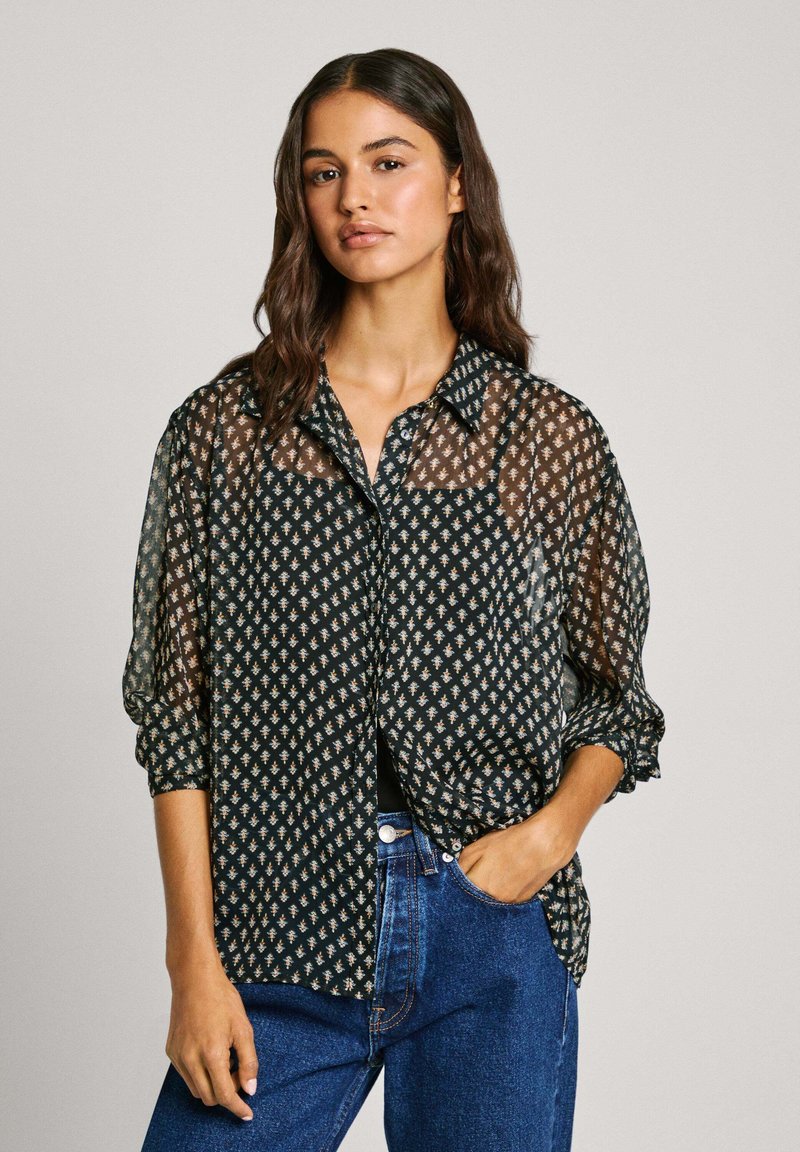 Durchsichtige schwarze Bluse mit einem diamantförmigen Muster in Beige, langen Ärmeln mit Knopfmanschetten, einer Brusttasche und lockerer Passform. Kombiniert mit blauen Jeans.