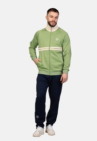 Grüne Zip-Jacke mit weißen Streifen, kombiniert mit marineblauen Hosen. Die Jacke hat einen hohen Kragen und ein weiches Material, mit einem Logo auf der Brust.