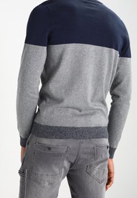 Maglione grigio e blu navy, con un design a blocchi di colore. Il tessuto ha una texture liscia con polsini e orlo a coste. Abbinato a pantaloni grigi.