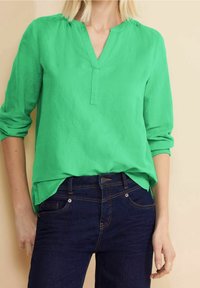 Grön, lätt blus med V-ringning och trekvartslånga ärmar, kombinerad med mörkblå denimjeans. Tyget har en mjuk och slät textur.