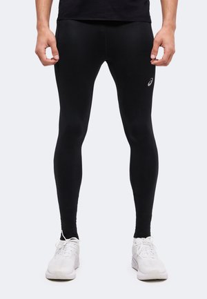 Homme portant un legging de sport noir ajusté et des chaussures de sport blanches, debout avec les mains détendues le long du corps devant un fond uni.