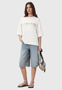 T-shirt biancooversized in cotone con testo "ALLSAINTS", abbinato a pantaloni corti in denim a vita alta con orli sfrangiati e sandali argentati. Borsa metallica inclusa.