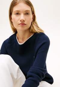 Ναυτικό πουλόβερ πλεκτό με στρογγυλή λαιμόκοψη και ριμπ υφή. Λευκό πουκάμισο από μέσα, συνδυασμένο με παντελόνι από υφασμάτινο ύφασμα ανοιχτόχρωμο.