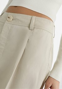 Detailní záběr na osobu v béžových kalhotách s vysokým pasem, které mají knoflík a poutka na pásek, doplněné bílým žebrovaným topem s dlouhým rukávem.