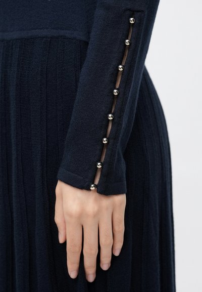 Ręka spoczywająca obok, ubrana w granatową prążkowaną sukienkę z przejrzystym paskiem i srebrnymi guzikami w kształcie kul wzdłuż mankietu rękawa.