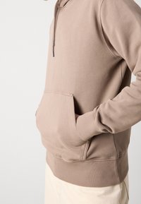 Person som bär en beige huvtröja med händerna i framfickan, i kombination med ljusa byxor, mot en vanlig vit bakgrund.