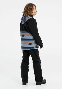 Schwarze und gemusterte Jacke mit einem blauen, braunen und schwarzen geometrischen Design. Das Model trägt schwarze Hosen und schwarze Stiefel, die die Kapuze der Jacke zur Geltung bringen.