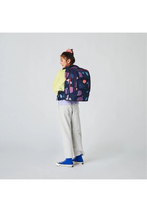 Young girl smiling, standing sideways wearing a navy backpack with colorful abstract patterns, light pants, yellow-green jacket, and blue sneakers.