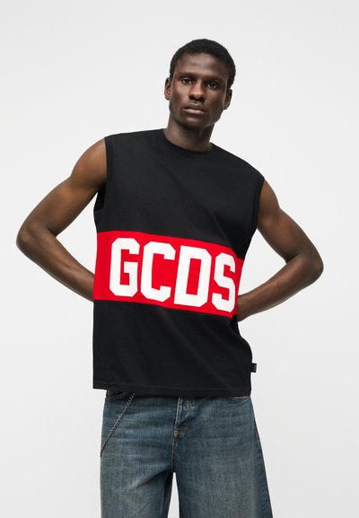 Άνδρας που φοράει αμάνικο μαύρο μπλουζάκι με φαρδιά κόκκινη ρίγα και μεγάλα λευκά γράμματα "GCDS", σε συνδυασμό με μπλε τζιν, στέκεται με τα χέρια στη μέση.