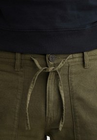 Pantalon vert olive avec un bouton et une ceinture en tissu nouée, associé à un haut noir à ourlet côtelé.