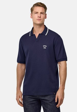 Tamsiai mėlynas polo marškinėliai su V formos iškirpte, balta apvado juosta ant apykaklės ir siuvinėtu logotipu ant krūtinės. Trumpomis rankovėmis ir tiesiu sijonu.