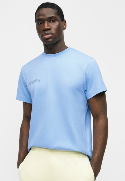 Άνδρας που φοράει ανοιχτό μπλε κοντομάνικο μπλουζάκι με μικρό τυπωμένο κείμενο στο στήθος και ανοιχτό κίτρινο παντελόνι, στέκεται με τα χέρια στις τσέπες.