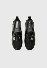 Schwarze Lederloafer mit runder Zehenform, silbernen Hardware-Akzenten und Schnürdetails. Das Markenlabel ist auf den Innensohlen sichtbar.