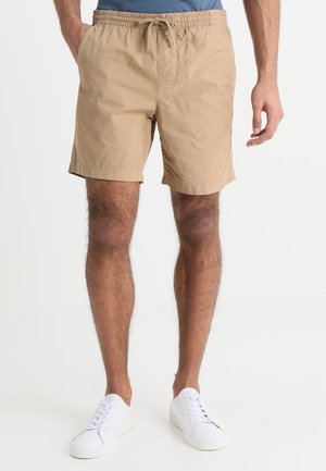 Man draagt beige short met trekkoord en witte sneakers, staand tegen een effen witte achtergrond met ontspannen handen langs het lichaam.