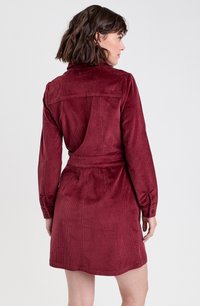 Robe en velours côtelé bordeaux avec un col, des manches longues et une taille ceinturée. Présente des nervures verticales et des poignets à boutons.