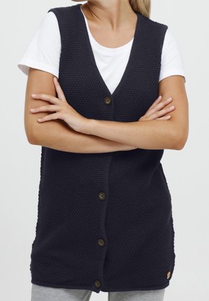 Persoon draagt een wit T-shirt met korte mouwen en een getextureerd marineblauw knoopjesloze mouwloos vest, met armen gekruist.