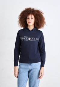 Hanorac bleumarin cu un grafic frontal care include "GANT 1949" și un scut. Material moale, mâneci lungi și glugă cu șnur. Asortat cu blugi deschiși.