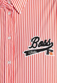 Chemise rayée verticale rouge et blanche avec des boutons blancs et un patch noir et blanc sur la poitrine indiquant "Boss Athletic".