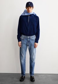 Marineblauwe knoopshirt, lichtblauwe sjaal, lichtgewassen rechte jeans, zwarte loafers en marineblauw pet. Glad stof, casual pasvorm, geen patronen.