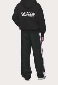 Felpa nera con testo "PEGADOR Next Wave" in rosa sulla schiena, abbinata a pantaloni sportivi neri con una striscia rosa laterale.