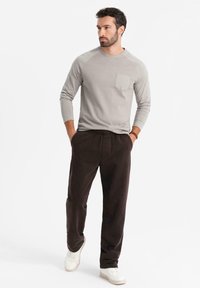 Maglietta a maniche lunghe grigia chiara con taschino sul petto e maniche raglan, abbinata a pantaloni marrone scuro e sneakers bianche.