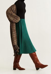 Falda de ante verde con una blusa negra ajustada, complementada con una bufanda marrón de gran tamaño con estampado y botas marrones hasta la rodilla.