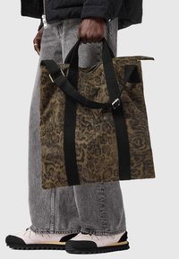 Borsa tote in tela con stampa leopardata marrone e manici neri, dotata di chiusura con zip e tasca esterna. Indossata con jeans grigi e scarpe chiare.