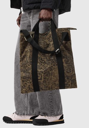 Sac en toile avec un imprimé léopard marron et des poignées noires, doté d'une fermeture zippée en haut et d'une poche extérieure. Porté avec un jean gris et des chaussures claires.
