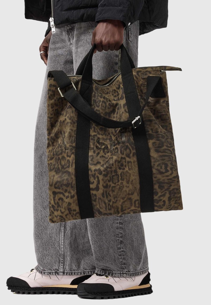 Canvas toteväska i brun leopardmönster med svarta remmar, med dragkedja upptill och ytterficka. Bärs med grå jeans och ljusa skor.