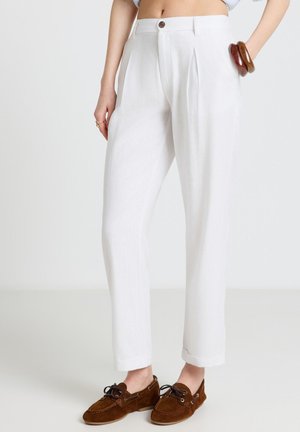 Donna che indossa pantaloni bianchi plissettati a vita alta con polsini, mocassini marroni in camoscio e braccialetti di legno al polso, in piedi contro uno sfondo semplice.