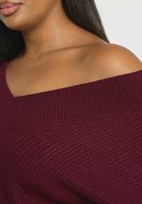 Burgundowy sweter bez ramiączek o żebrowanej fakturze i szerokim dekolcie. Wykonany z miękkiego materiału dzianinowego, z ukośnymi wzorami na powierzchni.